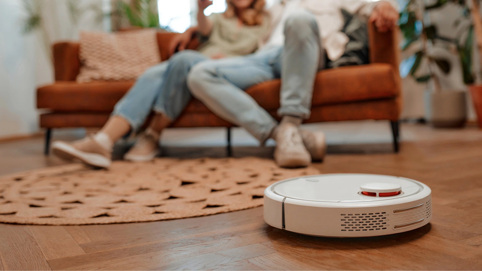 Salon zemininde çalışan akıllı robot süpürge, arkada koltukta oturan çift kapak görseli